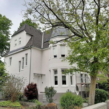 Weißes zweistöckiges Haus mit einem Turm und dunkelgrauem Dach. Bäume und Sträucher umgeben das Gebäude.
