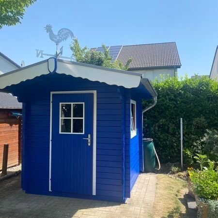 Blauer Gartenschuppen aus Holz mit weißen Zierleisten, einer Wetterfahne und einem kleinen Fenster.