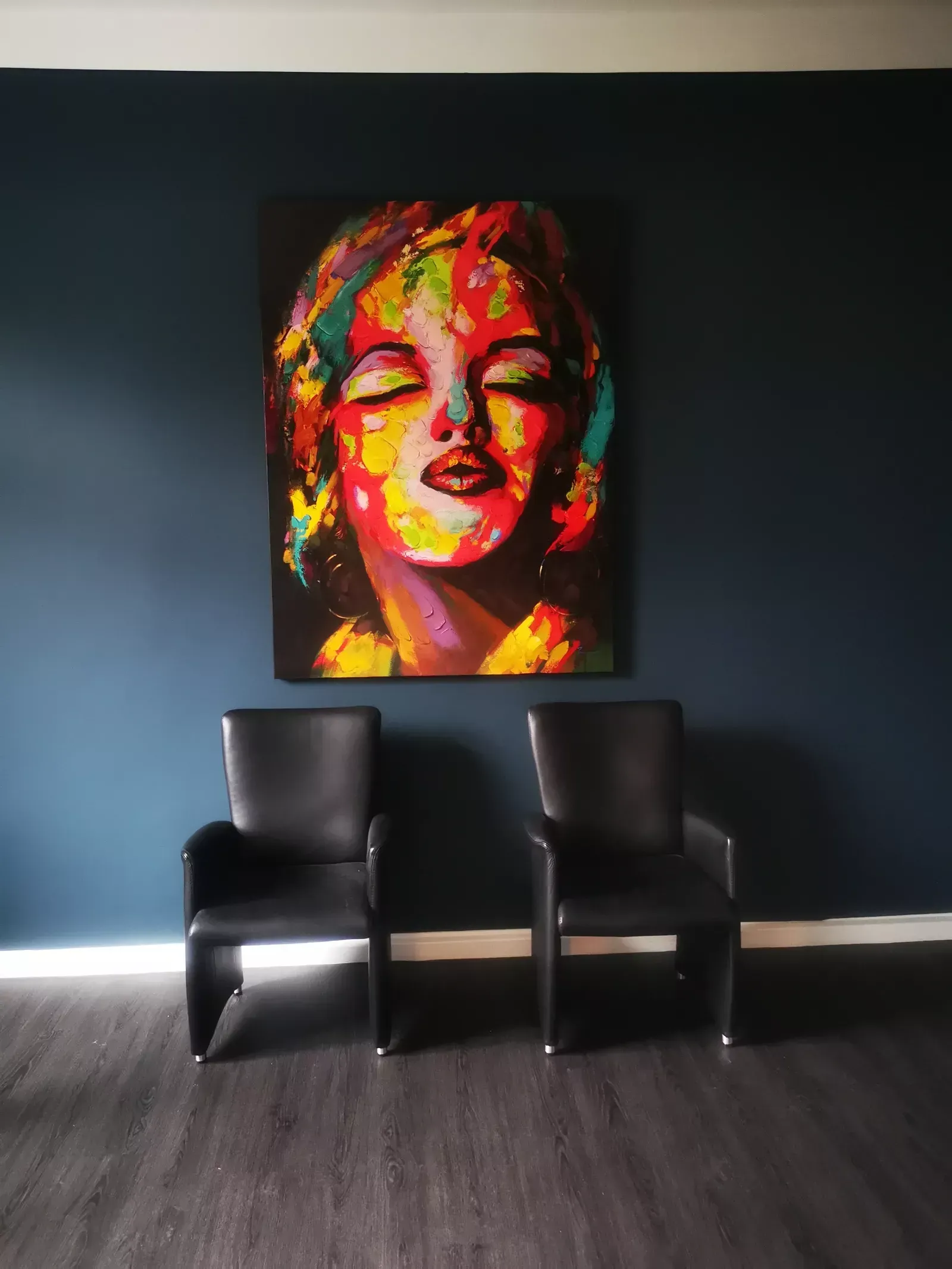 Ein Marilyn-Monroe-Gemälde hängt an einer dunkelblauen Wand über zwei schwarzen Stühlen in einem Raum mit Holzboden.