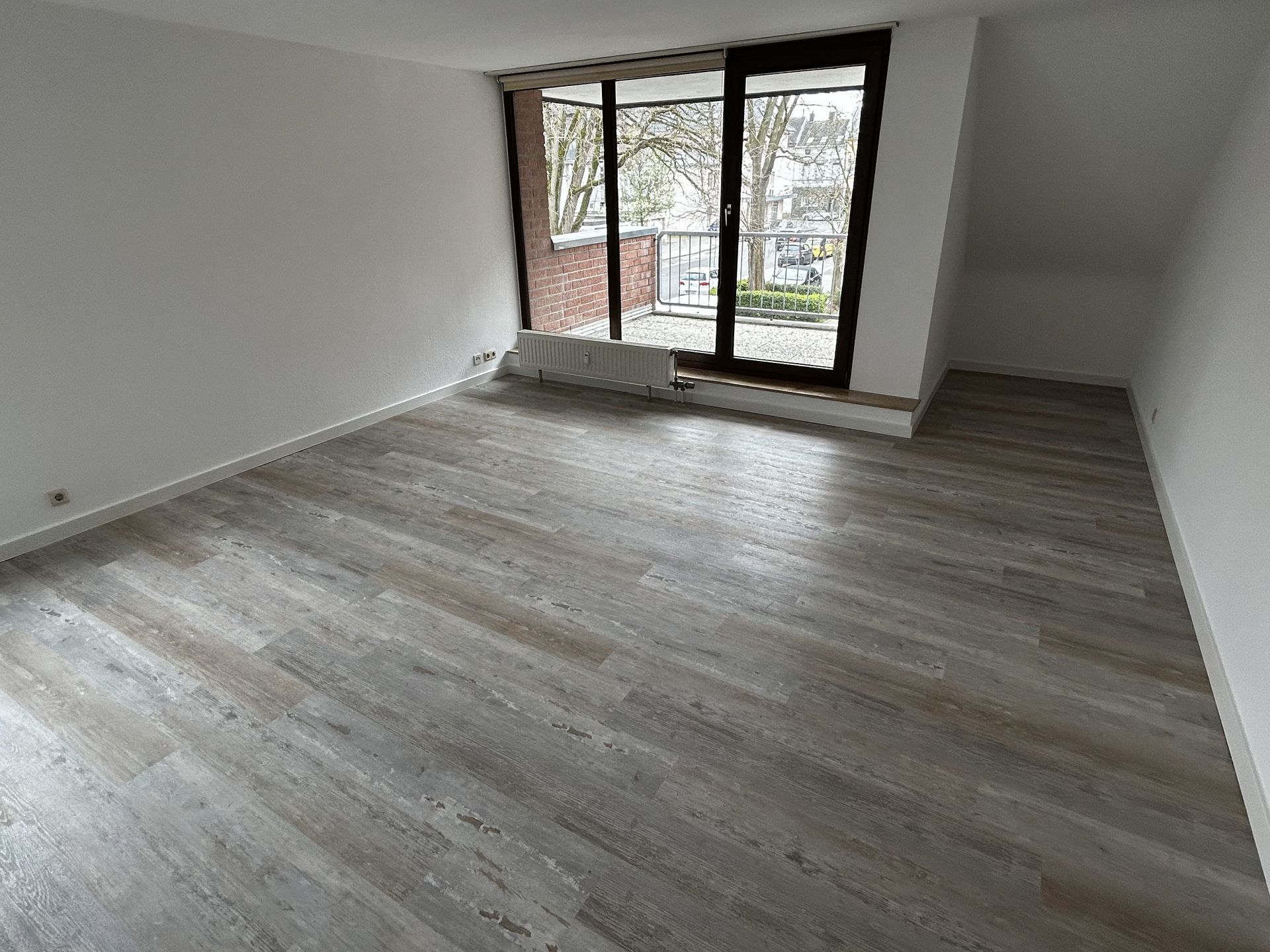 Leeres Zimmer mit Holzbodenoptik und einer Glasschiebetür, die auf einen Balkon führt. Weiße Wände.