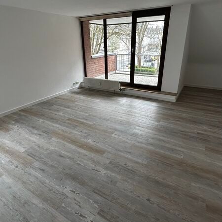 Leeres Zimmer mit Holzbodenoptik und Balkontür. Weiße Wände, natürliches Licht.