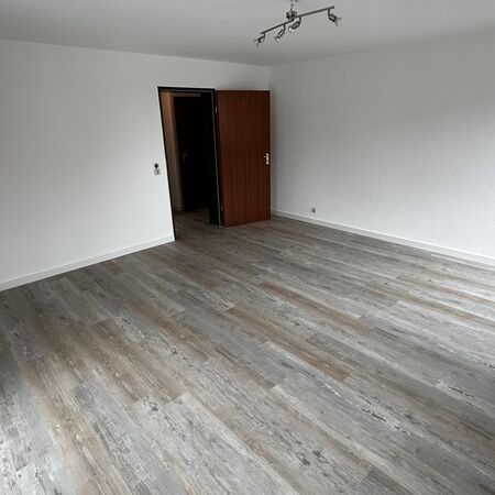 Ein leerer Raum mit Holzbodenoptik, weißen Wänden und einer geschlossenen braunen Tür, die zu einem dunklen Flur führt.