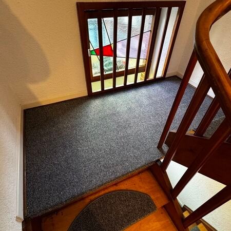 Treppenhaus mit Buntglasfenster, grauem Teppichboden und Holzgeländer.
