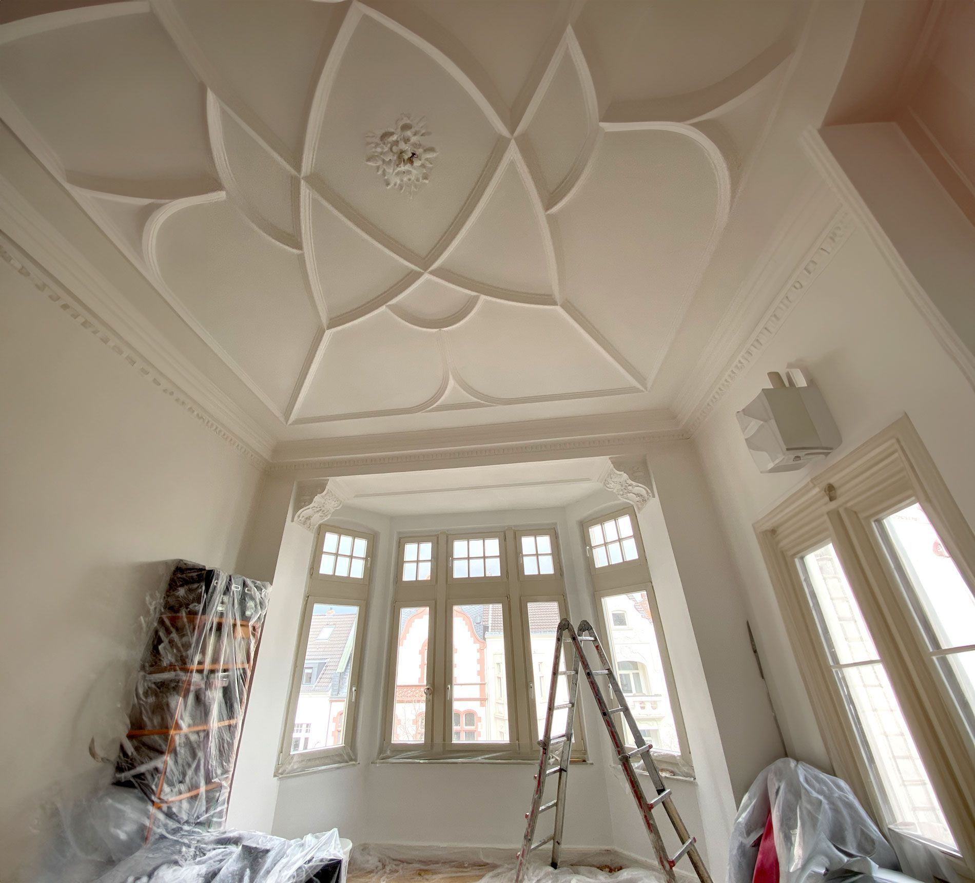 Verzierte weiße Decke mit floralem Muster, Erkerfenster, Leiter und eine teilweise bedeckte Wand.