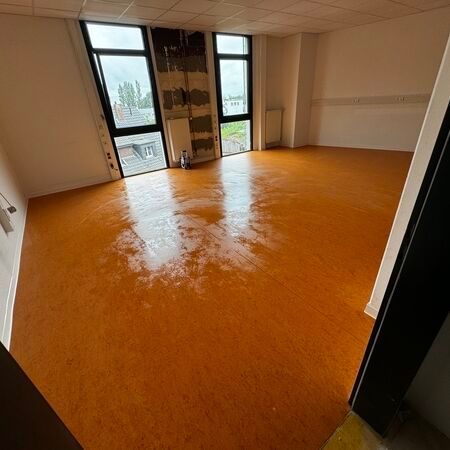 Leerer Raum mit orangefarbenem Boden, hohen Fenstern und weißen Wänden.