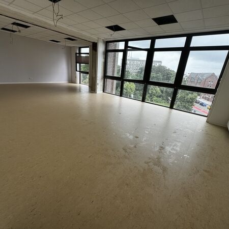 Leeres Zimmer mit großen Fenstern mit Blick auf die Stadt, hellem Fußboden und grauen Wänden.