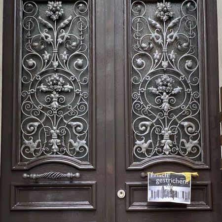 Verzierte, dunkelbraune Doppeltüren mit silbernen Schmiedeeisenverzierungen; an der rechten Tür ist ein Plakat angeklebt.