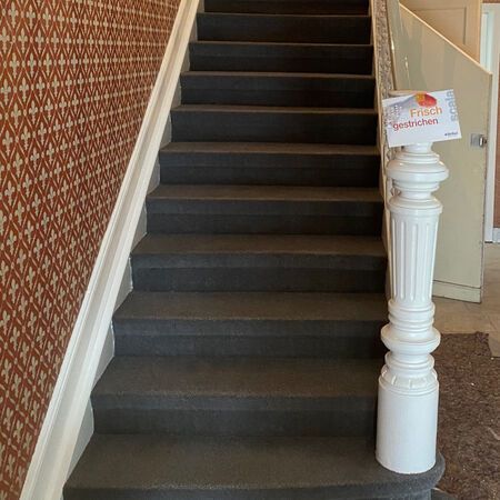 Treppe mit dunkelgrauen Stufen und gemusterter Tapete, weißem Treppengeländer und einem Schild an einem Pfosten.