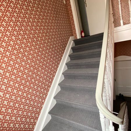 Treppenhaus mit grauem Teppichboden und weißen Zierleisten. Rote gemusterte Tapete und weißes Treppengeländer.
