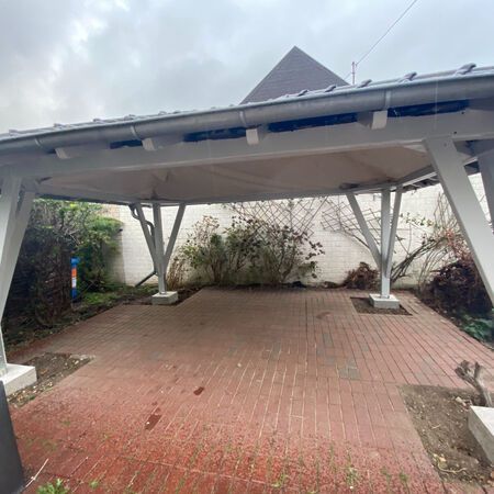 Carport mit rotem Backsteinboden, weißen Säulen und hellem Dach, neben einigen Büschen.