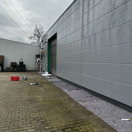 Arbeiter streichen die Außenfassade eines grauen Industriegebäudes; sie stehen auf Leitern.