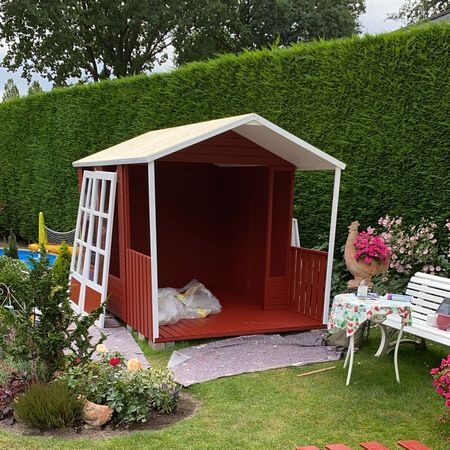 Ein rot-weißes Spielhaus in einem Garten mit Heckenhintergrund.
