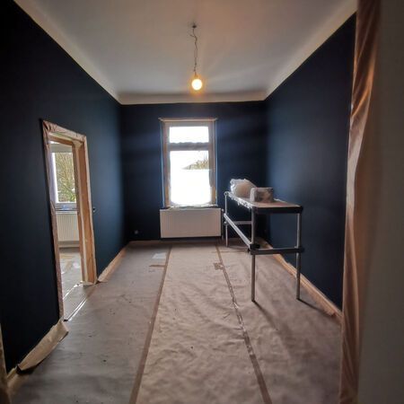 Zimmer mit dunkelblauen Wänden, weißer Decke, Fenster und einem provisorischen Arbeitstisch; Malerarbeiten im Gange.