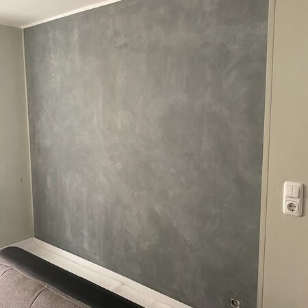 Graue, strukturierte Wand mit weißen Zierleisten, ein Sofa ist teilweise sichtbar.