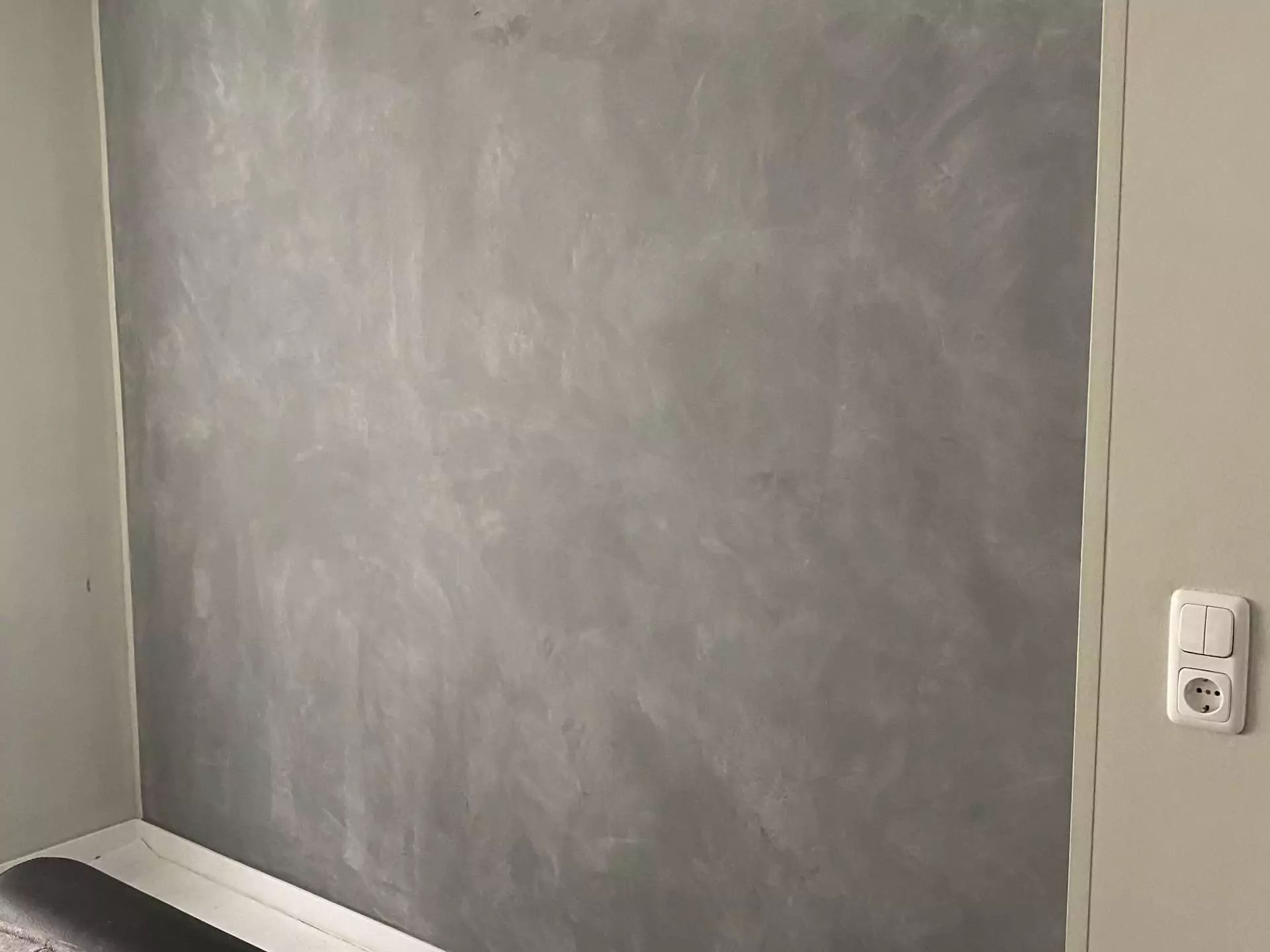 Graue Wand mit strukturierter Oberfläche, Zimmerecke, Steckdose rechts.
