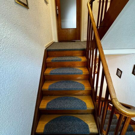 Holztreppe mit grauen Teppichstufen, die zu einer Tür führt; rechts ein Holzgeländer.