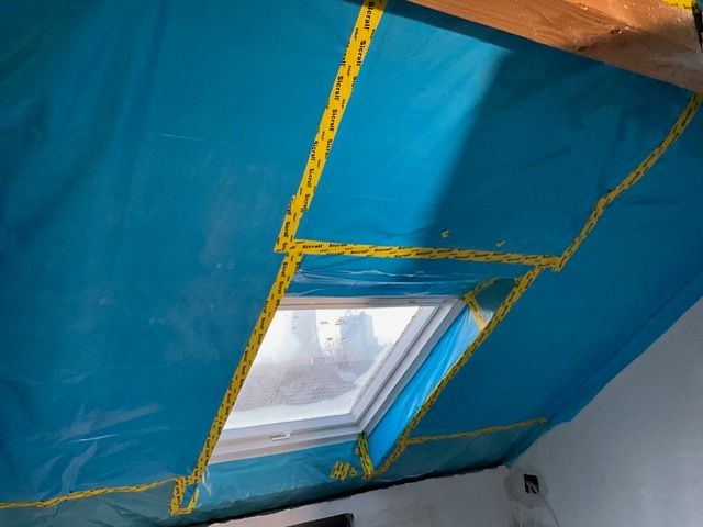 Blaue, isolierte Dachbodendecke mit einem kleinen, mit gelbem Klebeband umrahmten Dachfenster.