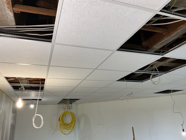 Beschädigte abgehängte Decke mit fehlenden Platten, freiliegenden Kabeln und herabhängenden Lampen in einem Zimmer