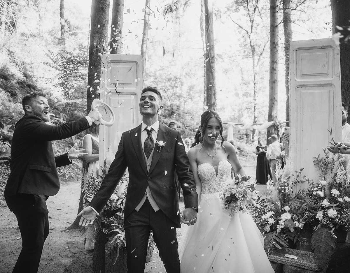 Una fotografía en blanco y negro de unos novios caminando por un bosque.