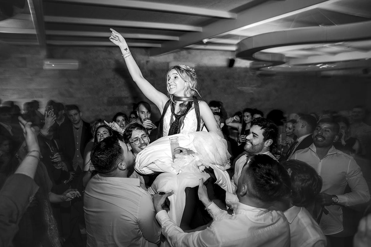 Una novia es levantada en el aire por un grupo de personas en una recepción de boda.
