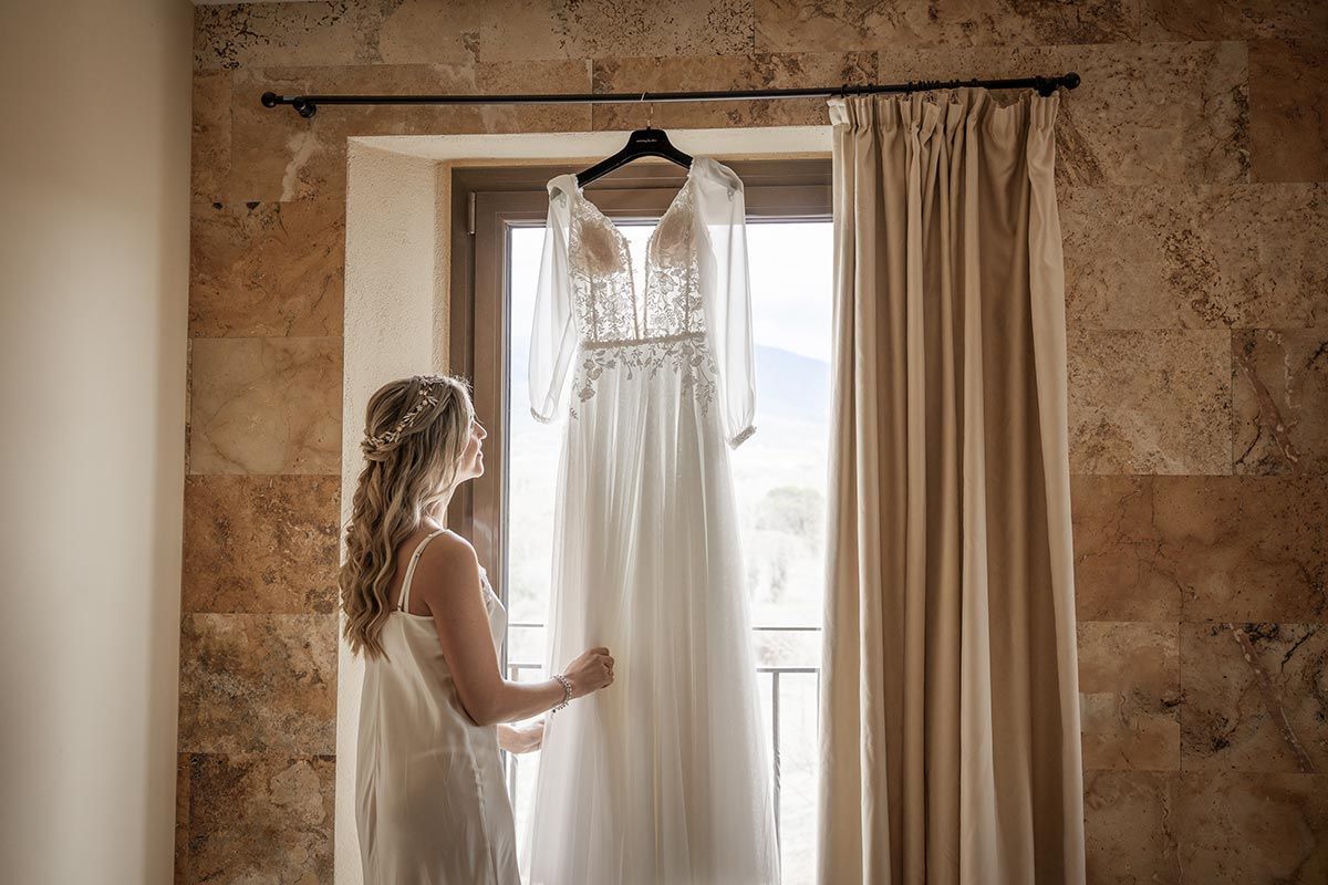 Una mujer mira desde una ventana su vestido de novia colgado en una percha.
