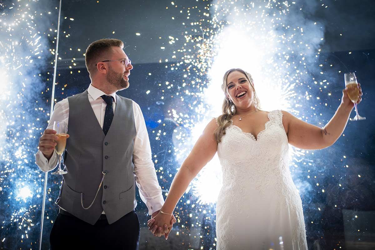 Una novia y un novio se toman de la mano y brindan con champán frente a los fuegos artificiales.