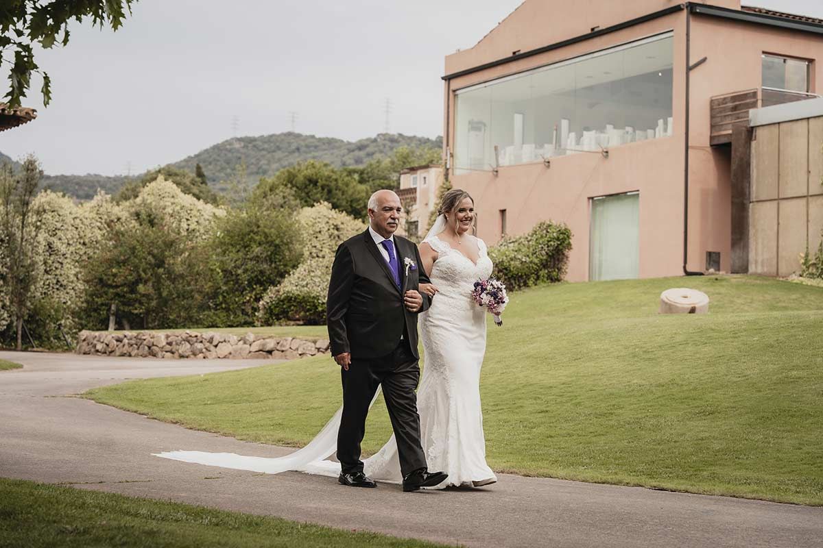 Una novia y su padre caminan por un sendero frente a un edificio.