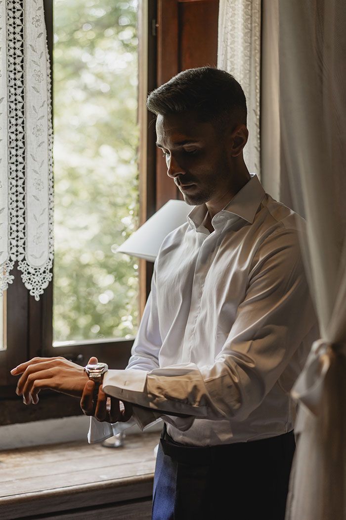 Un hombre con una camisa blanca mira su reloj mientras está parado frente a una ventana.