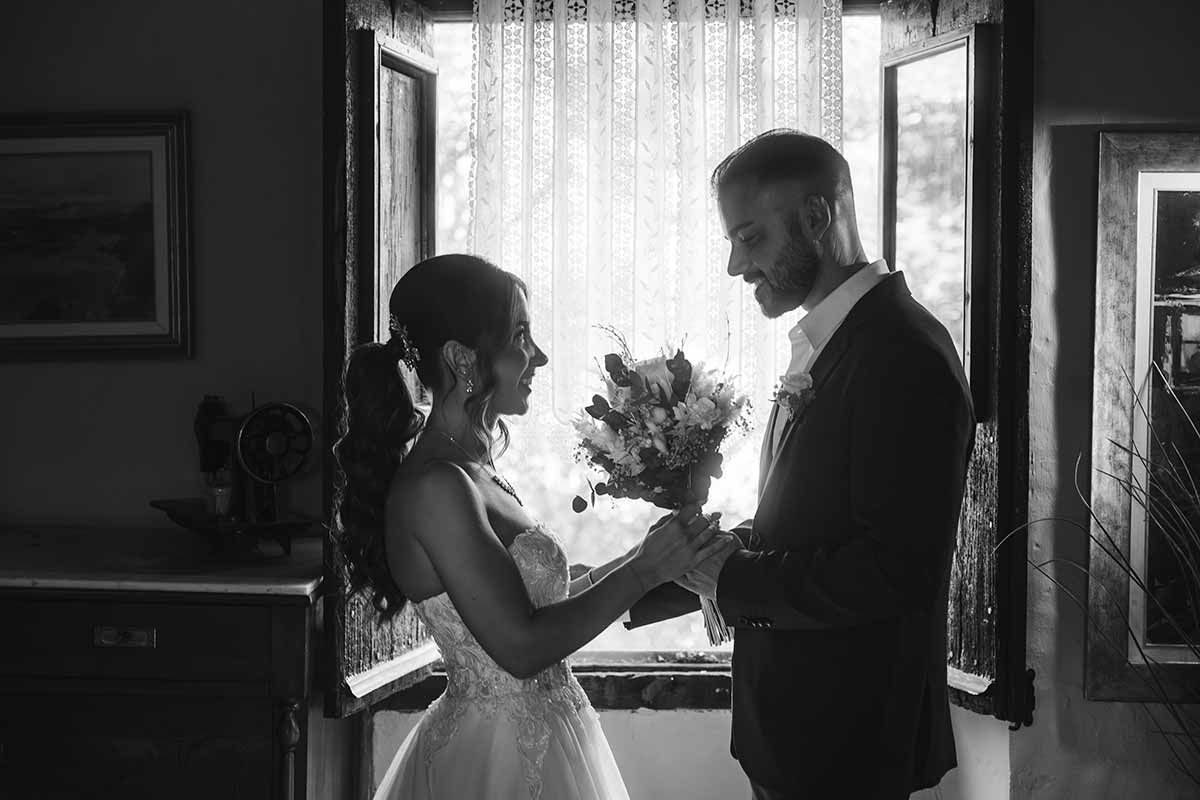 Una fotografía en blanco y negro de una novia y un novio tomados de la mano frente a una ventana.