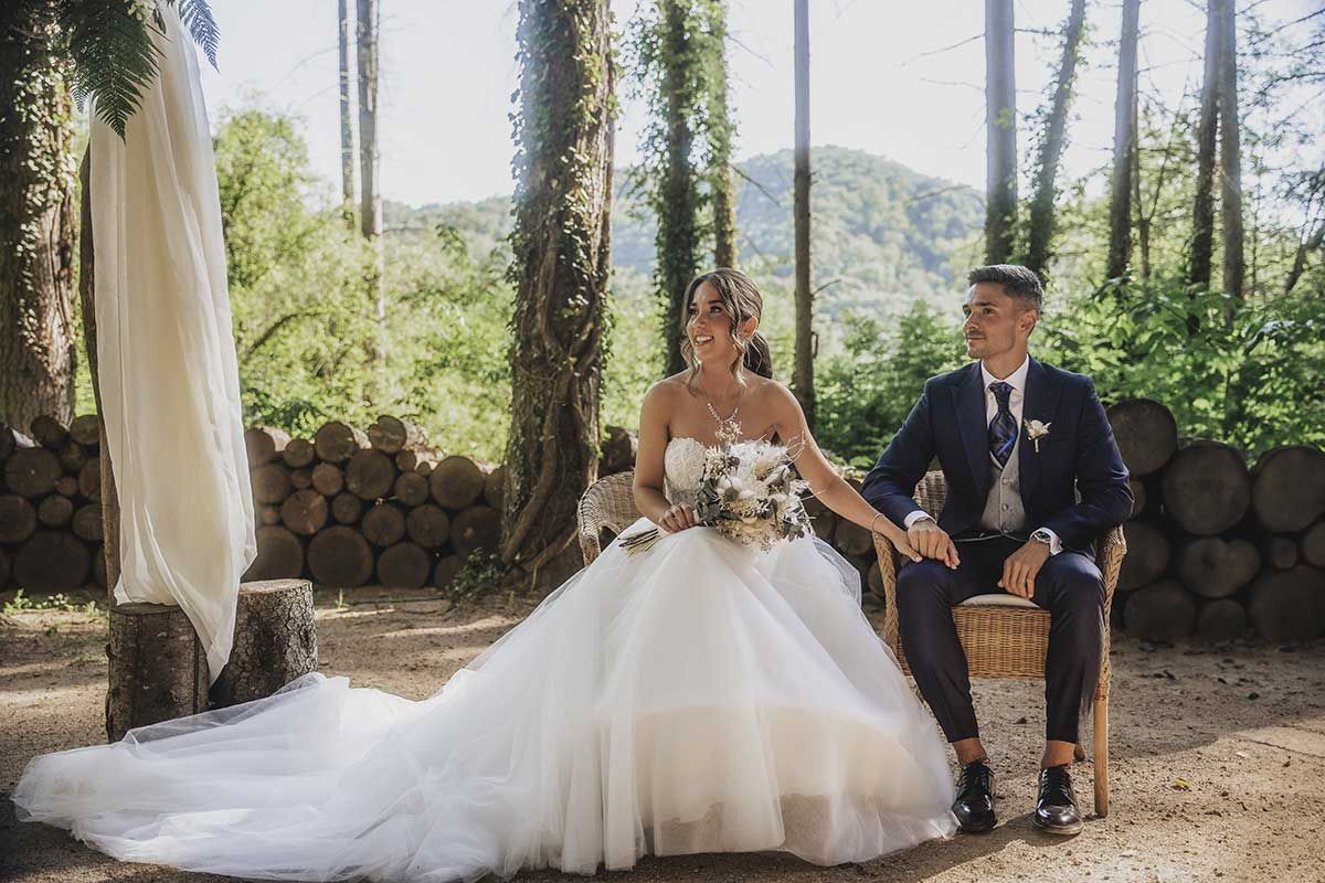 Una novia y un novio están sentados en sillas en el bosque tomados de la mano.