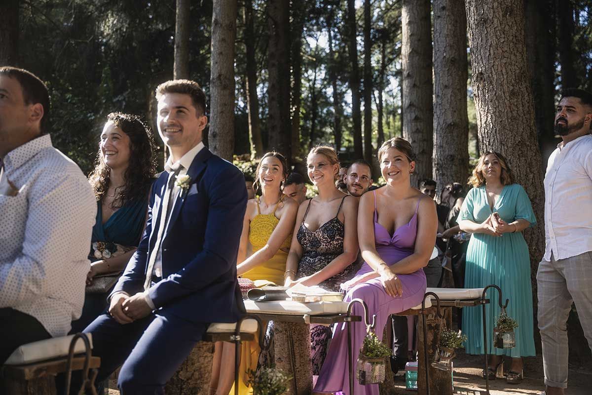 Un grupo de personas está sentada en un bosque viendo una ceremonia de boda.