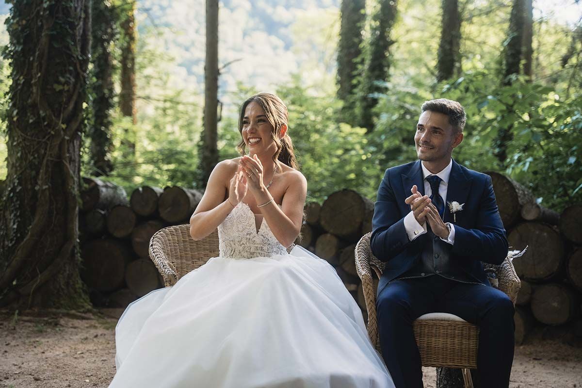 Una novia y un novio están sentados en sillas en el bosque aplaudiendo.