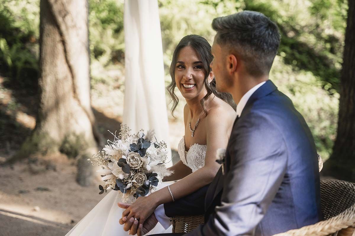 Una novia y un novio están sentados en una silla en el bosque tomados de la mano.