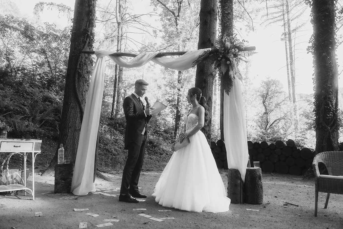 Una novia y un novio están de pie bajo un dosel en el bosque durante su ceremonia de boda.
