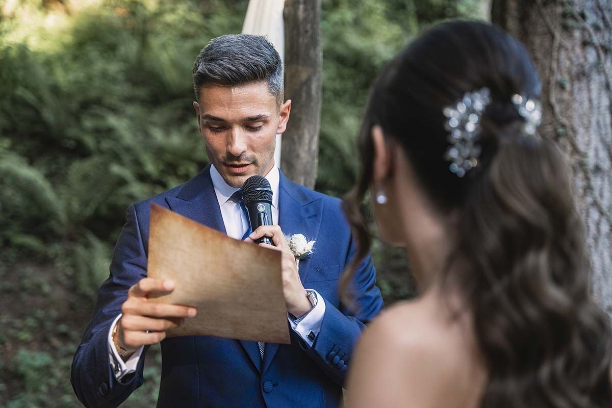 Un hombre con traje y corbata le lee un trozo de papel a una novia.