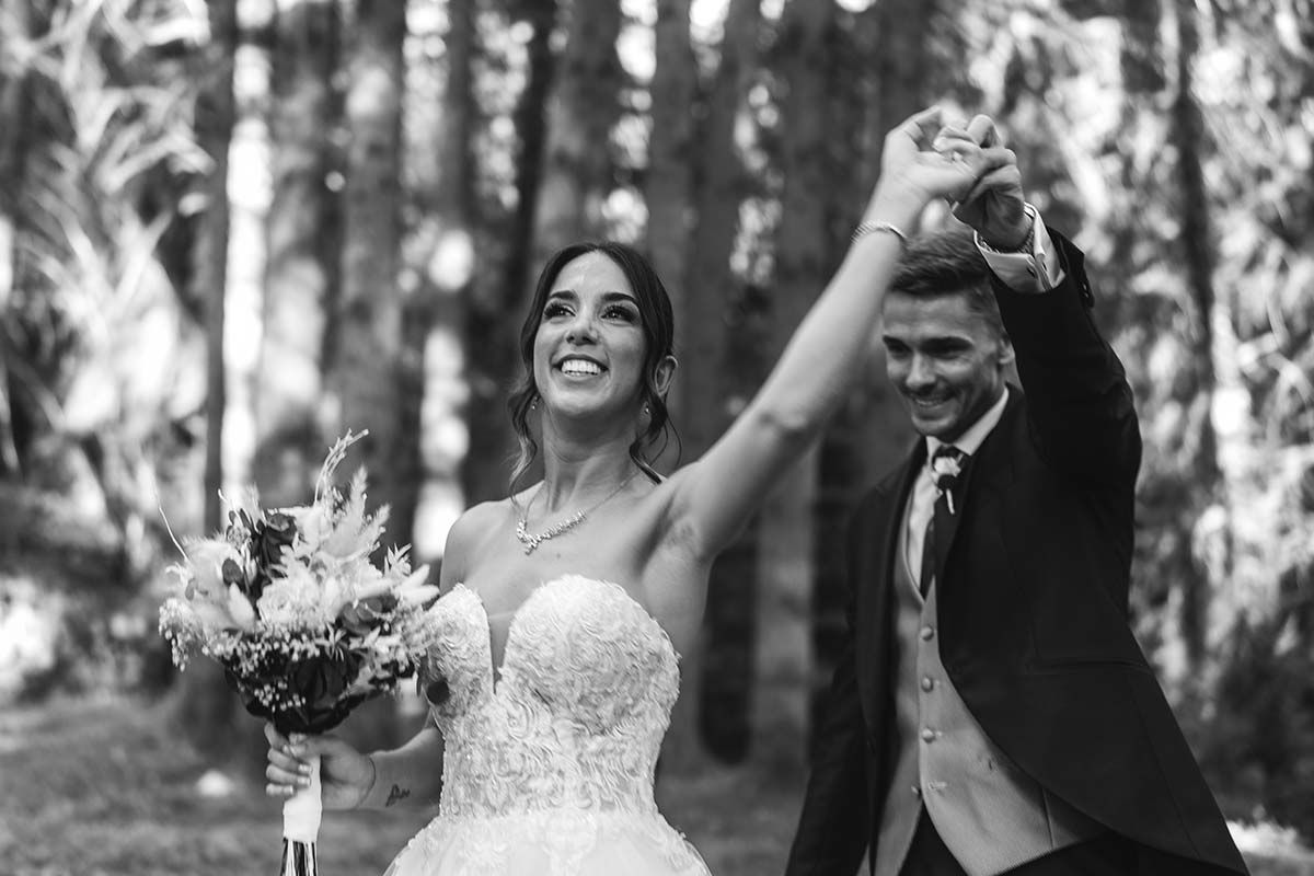 Una fotografía en blanco y negro de una novia y un novio tomados de la mano en el bosque.