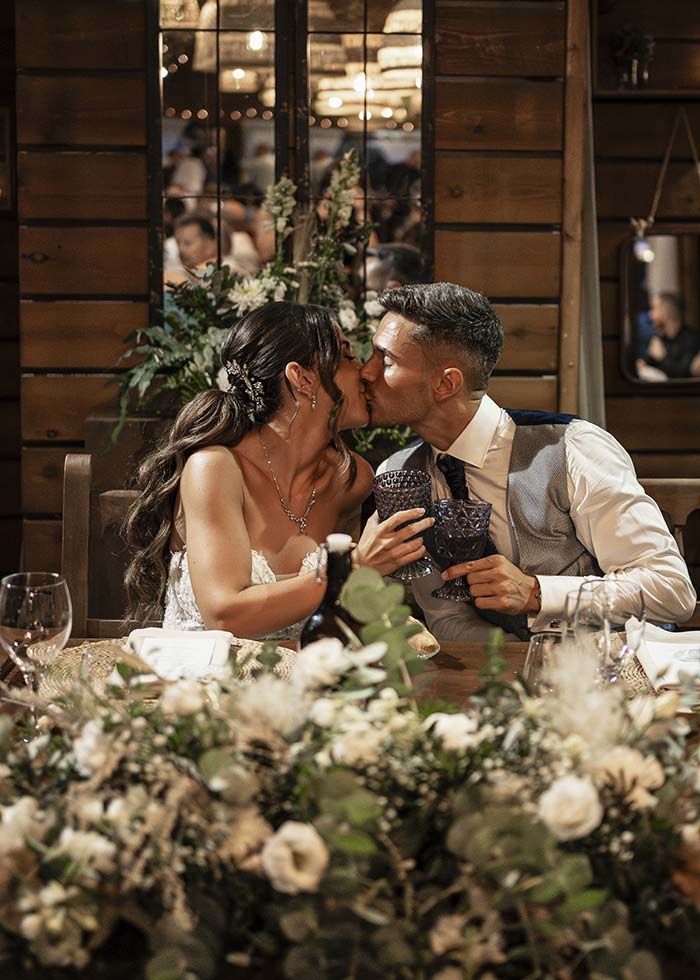 Una novia y un novio se besan en una mesa larga en la recepción de su boda.