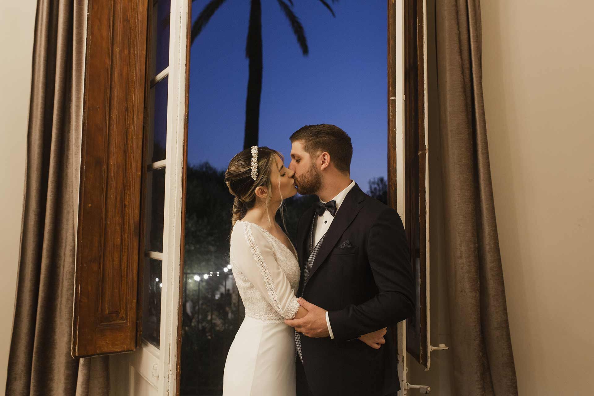 Una novia y un novio se besan delante de una ventana.