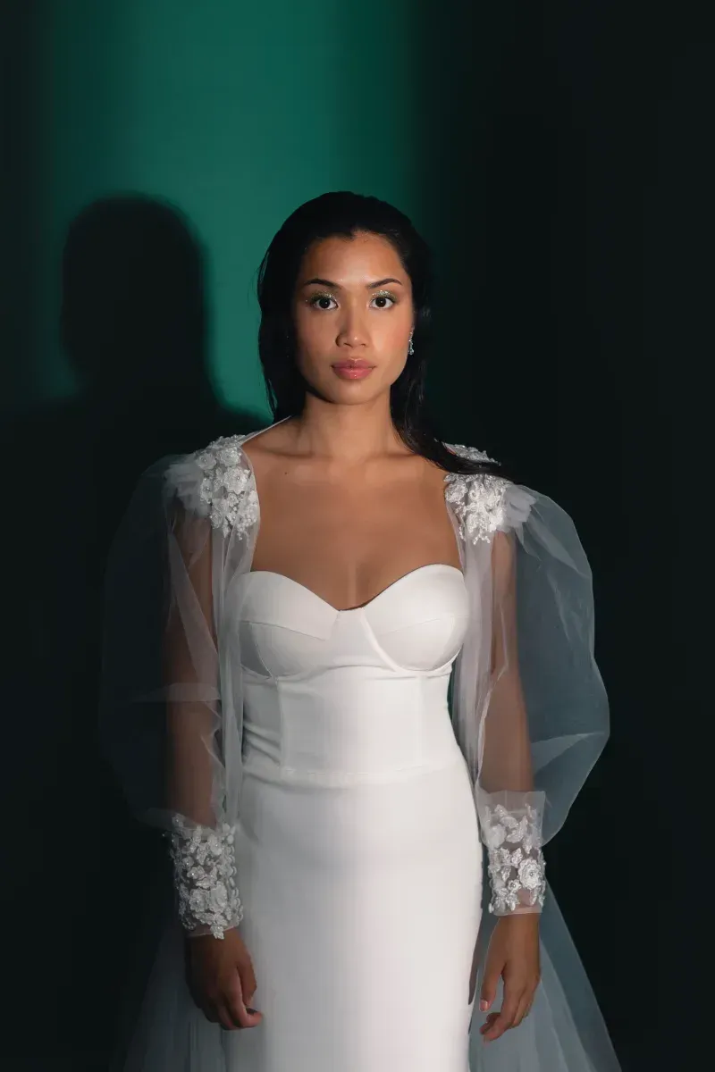 Una mujer con un vestido blanco con mangas abullonadas está parada frente a una pared verde.