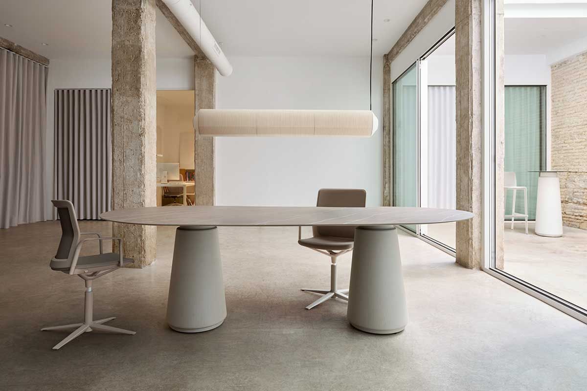 Bureau moderne avec table ovale, supports cylindriques, chaises et grandes fenêtres.