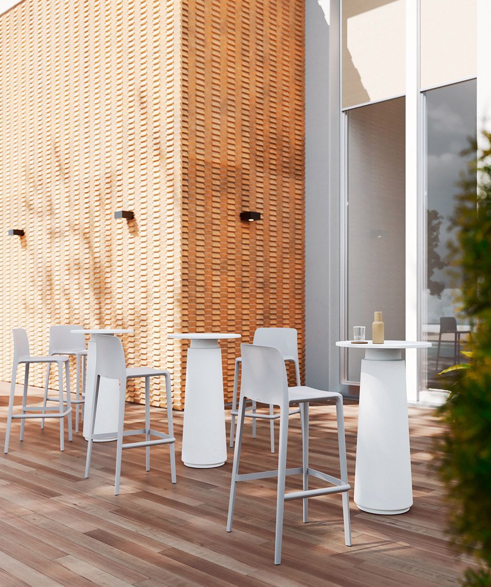 Terrasse extérieure avec tables de bar et tabourets blancs adossés à un mur de briques et une grande fenêtre.