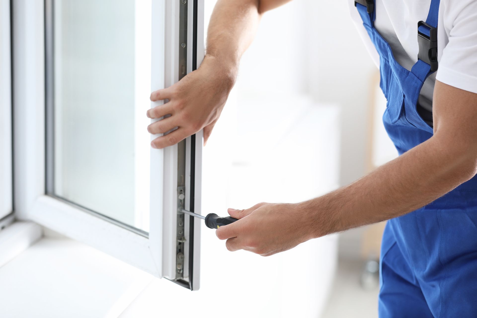 Mann im blauen Overall benutzt einen Schraubenzieher, um einen weissen Fensterrahmen einzubauen.