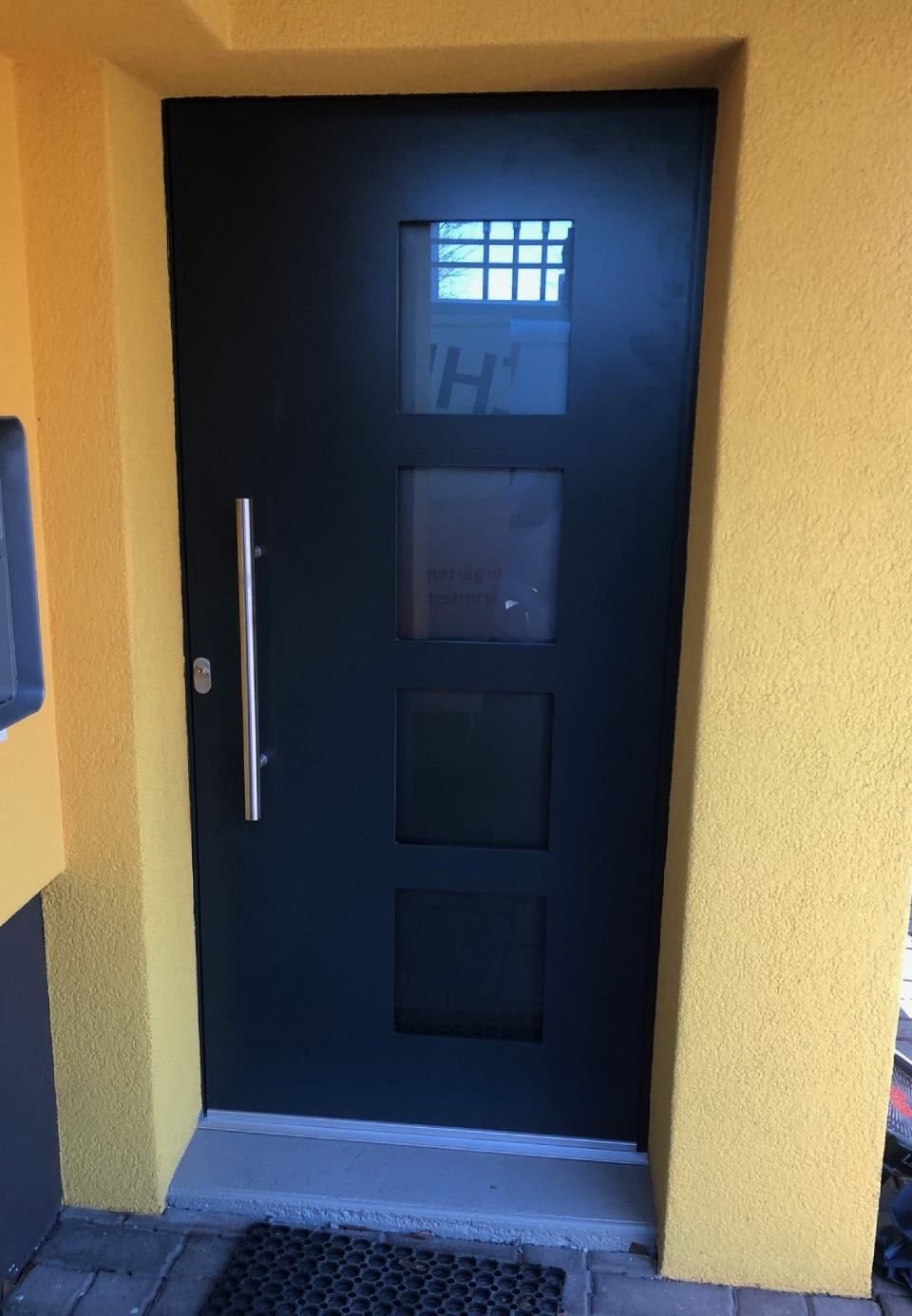 Schwarze Haustür mit drei quadratischen Fenstern und einem silbernen Griff in einer gelben Wand.