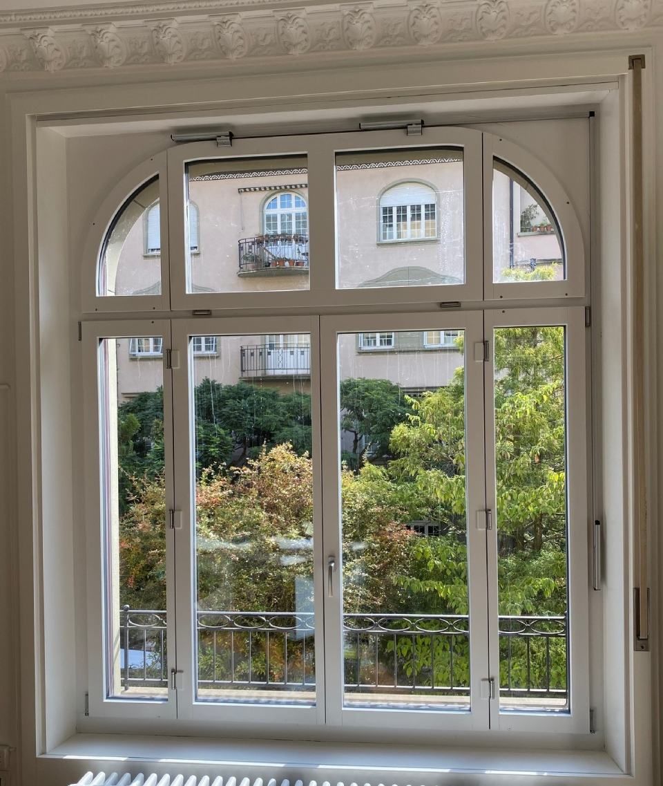 Fenster mit gewölbter Oberseite, geteilten Scheiben, mit Blick auf Bäume und ein Gebäude mit Balkonen. Weißer Rahmen.