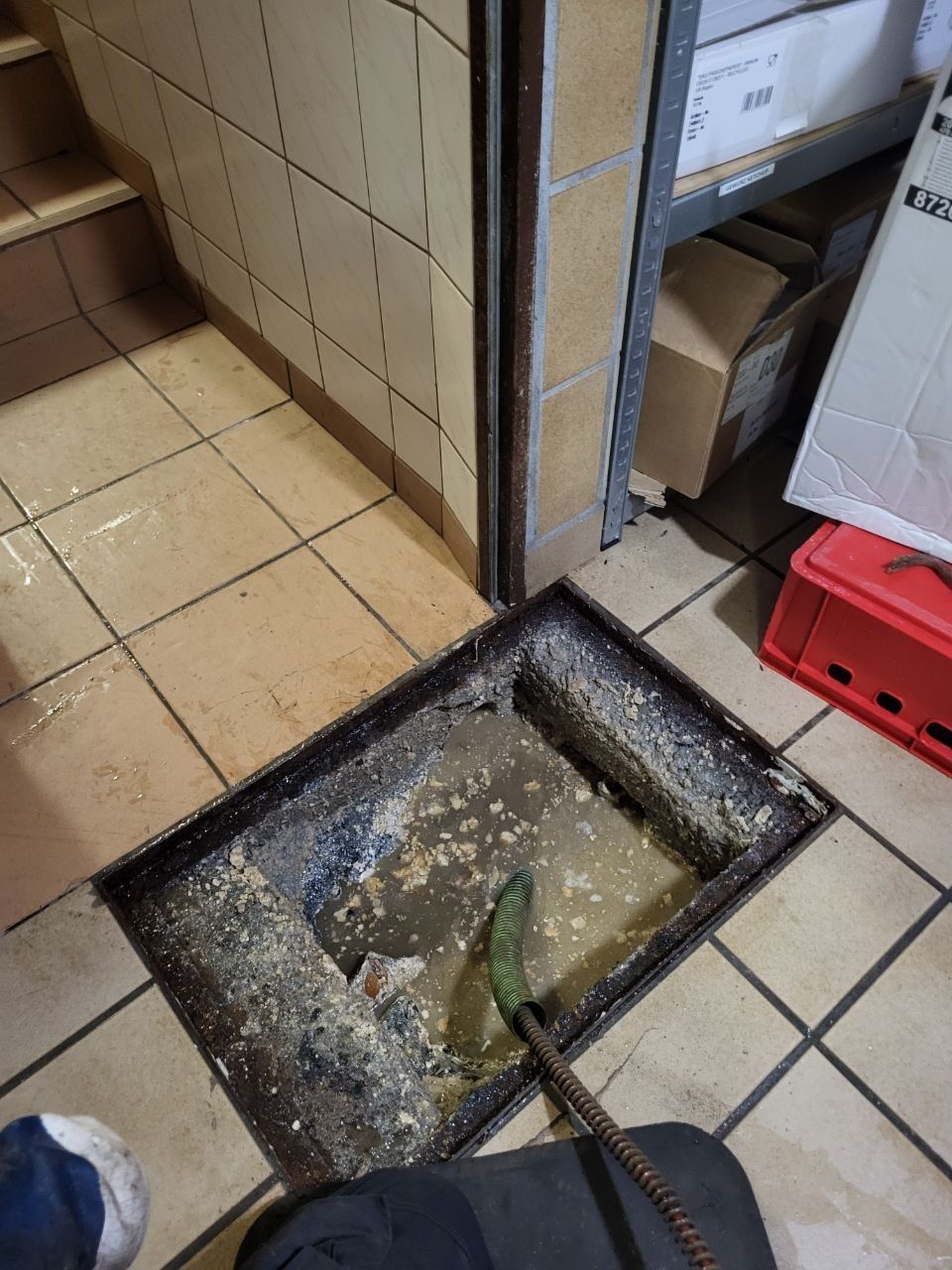 Ein Arbeiter in orangefarbener Sicherheitshose steht neben einem offenen Kanaldeckel auf nassem Betonboden.