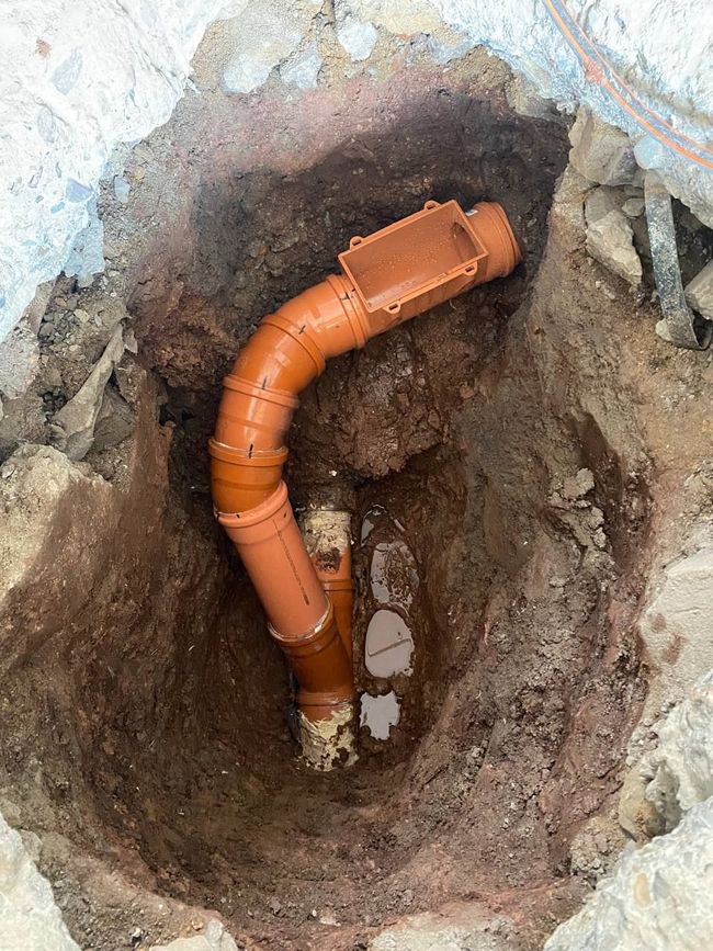 Versorgungsleitungen in einem Graben auf einer Baustelle, daneben Erdhaufen und blaue Zaunelemente.
