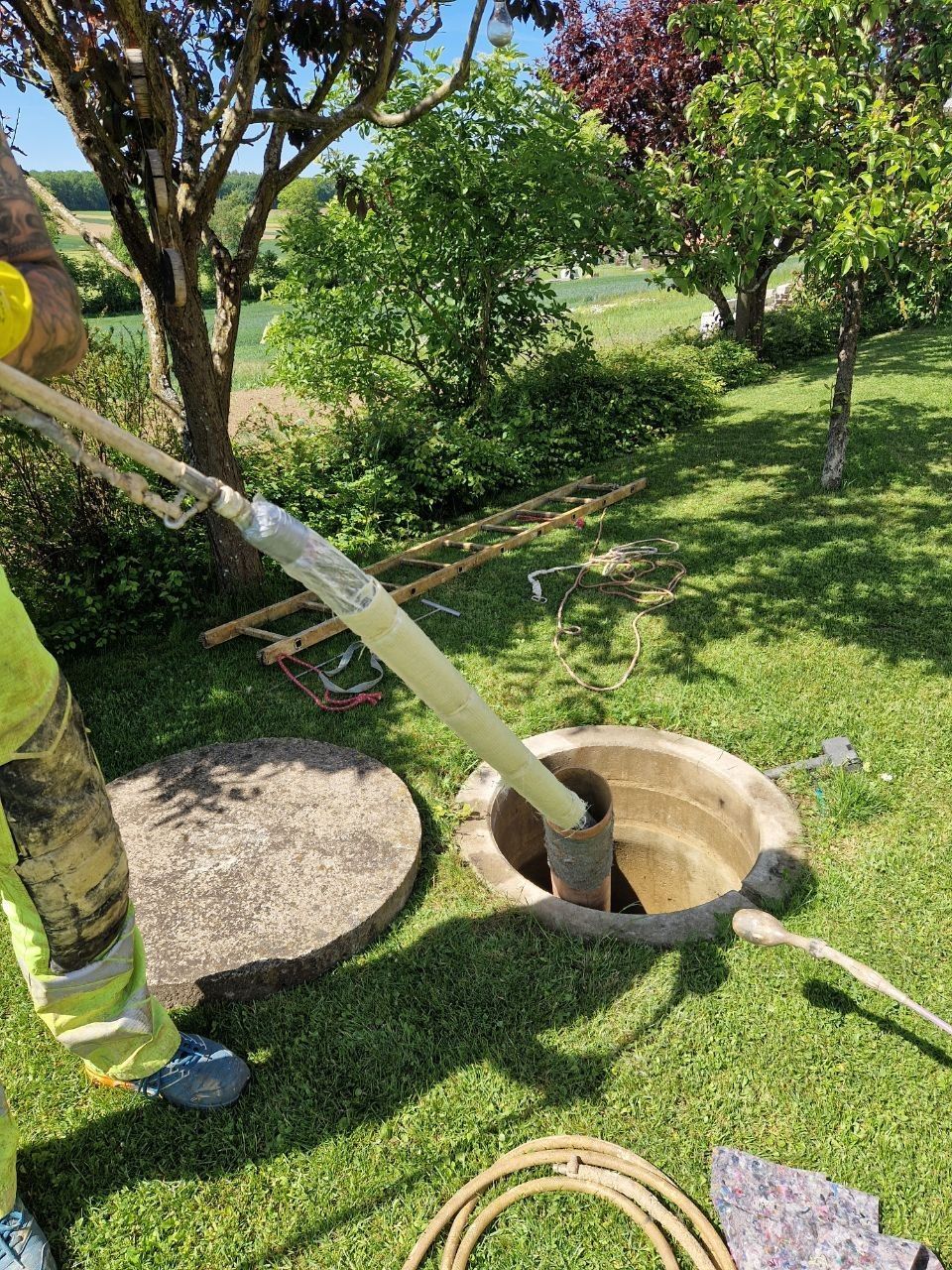 Ein Arbeiter saugt mit einem Schlauch durch einen offenen Gullydeckel einen grasbewachsenen Hof ab.