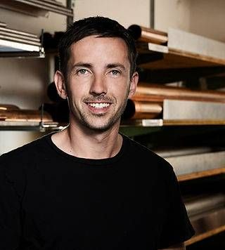 Eine Person in einem Hemd lächelt, während sie in einer Werkstatt steht; im Hintergrund sind Regale zu sehen.