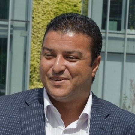 Um homem de terno e camisa branca está sorrindo em frente a um prédio.