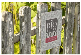 Ein Schild von Bio Terra Garten an einem verwitterten Holzzaun, dahinter grüne Pflanzen.